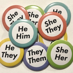 Könnte beinhalten: Neun runde Knöpfe in verschiedenen Farben mit Text. Der Text auf den Knöpfen lautet "She They", "He Him", "She Her", "He They", "They Them", "She" und "He".