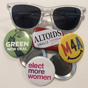 May include: Clear-framed sunglasses rest above a collection of buttons and a small Altoids tin. The buttons display the phrases "GREEN NEW DEAL", "M4A", and "elect more women". The Altoids tin is red and white.