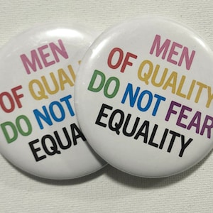May include: Two white button badges with the text "Men of Quality Do Not Fear Equality" in rainbow colours. The text is arranged in a circle around the centre of the badge.