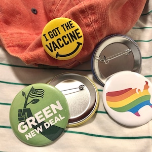 May include: A collection of round pinback buttons with various designs. One button reads "I GOT THE VACCINE" on a yellow background. Another features a rainbow-colored map of the United States. A third button says "GREEN NEW DEAL".