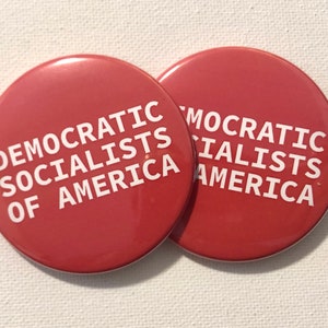 以下が含まれることがあります： 白い文字が書かれた赤い丸いボタンが2つ。ボタンの文字は「Democratic Socialists of America」です。