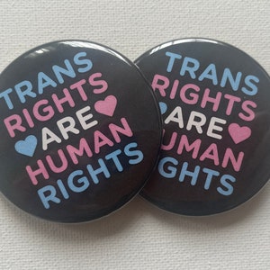 May include: Two black button badges with the text "Trans Rights Are Human Rights" in pink, blue, and white lettering. The text is arranged in a circle around a pink and blue heart.