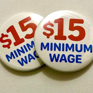 May include: Two white button pins with blue and red text. The text on the pins reads "$15 Minimum Wage".