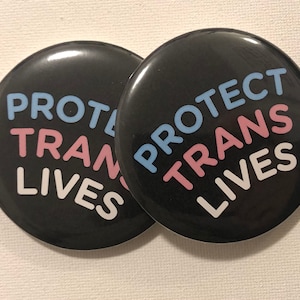 May include: Two black button pins with the text "PROTECT TRANS LIVES" in white, pink, and blue lettering.