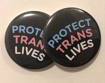 May include: Two black button pins with the text "PROTECT TRANS LIVES" in white, pink, and blue lettering.