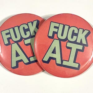 Puede incluir: Dos botones redondos rojos con el texto "FUCK AI" en letras verdes y azules. Los botones se muestran sobre una superficie blanca.