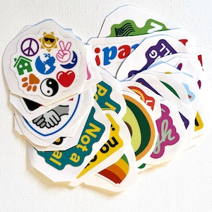 May include: A collection of colorful stickers with various designs and phrases, including peace signs, a globe, a house, a paw print, a yin yang symbol, a heart, a rainbow, an avocado, and the words "I am not a pro", "no", "peace", "laugh", and "I'll be back".