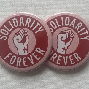 May include: Two pink round buttons with the text "Solidarity Forever" in white lettering. The buttons feature a raised red fist in the center.