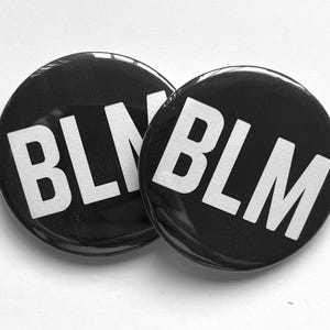May include: Two black button pins with white text that reads "BLM".