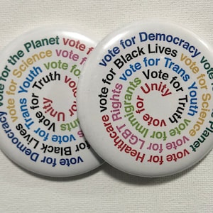 May include: Two white round buttons with colorful text. The buttons say "Vote for Democracy", "Vote for Black Lives", "Vote for Trans Rights", "Vote for Youth", "Vote for Science", and "Vote for the Planet".