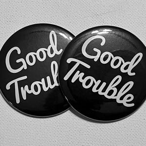 Könnte beinhalten: Zwei schwarze, runde Buttons mit dem Schriftzug "Good Trouble" in weißer Schreibschrift. Die Buttons haben eine glänzende Oberfläche. Der Hintergrund ist hell und neutral.