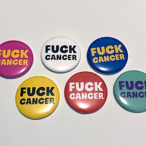 May include: A collection of round buttons in various colors, including pink, white, blue, yellow, red, and teal. Each button features the phrase "FUCK CANCER" in bold, contrasting colors.