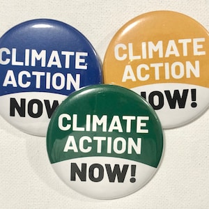 Könnte beinhalten: Drei runde Knöpfe mit weißem Text auf farbigem Hintergrund. Die Knöpfe zeigen "Climate Action Now!" in Blau, Gold und Grün.