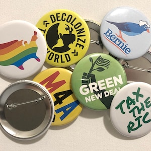 May include: A collection of round buttons with various designs and text. One button features a rainbow-colored map of the United States. Other buttons display phrases like "Decolonize World," "Bernie," "Green New Deal," and "Tax the Rich."