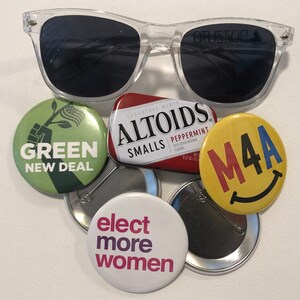 Puede incluir: Gafas de sol con montura transparente sobre una colecci&oacute;n de botones pol&iacute;ticos y una peque&ntilde;a lata de Altoids. Los botones muestran mensajes como "Green New Deal" y "Elect More Women". La lata de Altoids es roja y blanca.