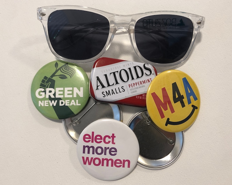 Puede incluir: Gafas de sol con montura transparente sobre una colecci&oacute;n de botones pol&iacute;ticos y una peque&ntilde;a lata de Altoids. Los botones muestran mensajes como "Green New Deal" y "Elect More Women". La lata de Altoids es roja y blanca.
