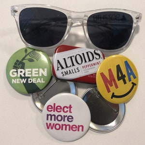May include: A collection of six round button pins with various political slogans. The pins are green, yellow, white, and red. The slogans include "Green New Deal", "M4A", "Elect More Women", and "Altoids Smalls Peppermint".