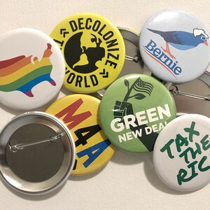 Puede incluir: Una colecci&oacute;n de botones redondos con varios dise&ntilde;os y texto. Un bot&oacute;n presenta un mapa de los Estados Unidos con los colores del arco&iacute;ris. Otros botones muestran frases como "Decolonize World" y "Tax the Rich."