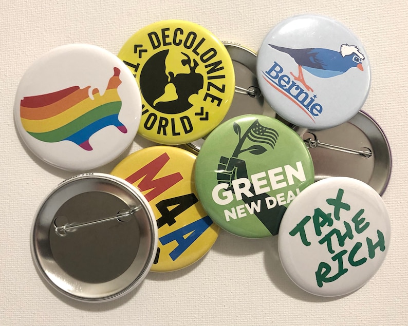 Puede incluir: Una colecci&oacute;n de botones redondos con varios dise&ntilde;os y texto. Un bot&oacute;n presenta un mapa de los Estados Unidos con los colores del arco&iacute;ris. Otros botones muestran frases como "Decolonize World" y "Tax the Rich."