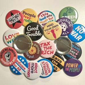 May include: A selection of circular badges with diverse political and social justice slogans. The badges display phrases such as "Dismantle the Oligarchy," "Good Trouble," and "Medicare for All," in various fonts and colours.