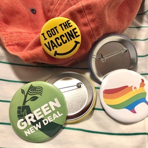 Puede incluir: Un conjunto de botones redondos con varios dise&ntilde;os. Uno dice "I GOT THE VACCINE" con una cara sonriente. Otro presenta un mapa de los Estados Unidos con los colores del arco&iacute;ris. Un tercer bot&oacute;n dice "GREEN NEW DEAL" con un gr&aacute;fico de una mano sosteniendo una planta.