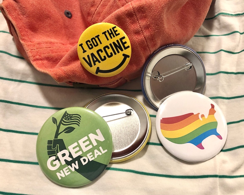 Puede incluir: Un conjunto de botones redondos con varios dise&ntilde;os. Uno dice "I GOT THE VACCINE" con una cara sonriente. Otro presenta un mapa de los Estados Unidos con los colores del arco&iacute;ris. Un tercer bot&oacute;n dice "GREEN NEW DEAL" con un gr&aacute;fico de una mano sosteniendo una planta.