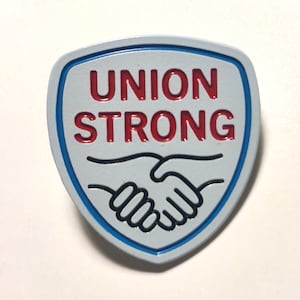 Könnte beinhalten: Eine blaue und weiße Emaille-Anstecknadel mit einem Händedruck-Design und dem Text "UNION STRONG" in roten Buchstaben.