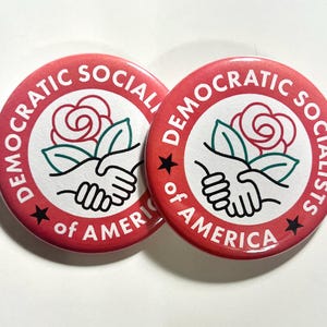 May include: Two red round buttons with the text "DEMOCRATIC SOCIALISTS of AMERICA" encircling a white center. The center features a red rose, green leaves, and clasped hands. Black stars are on the red border.