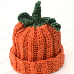 Op de afbeelding: Een gebreide pompoenmuts met een groene steel en bladeren. De muts is oranje en heeft een geribbelde textuur.