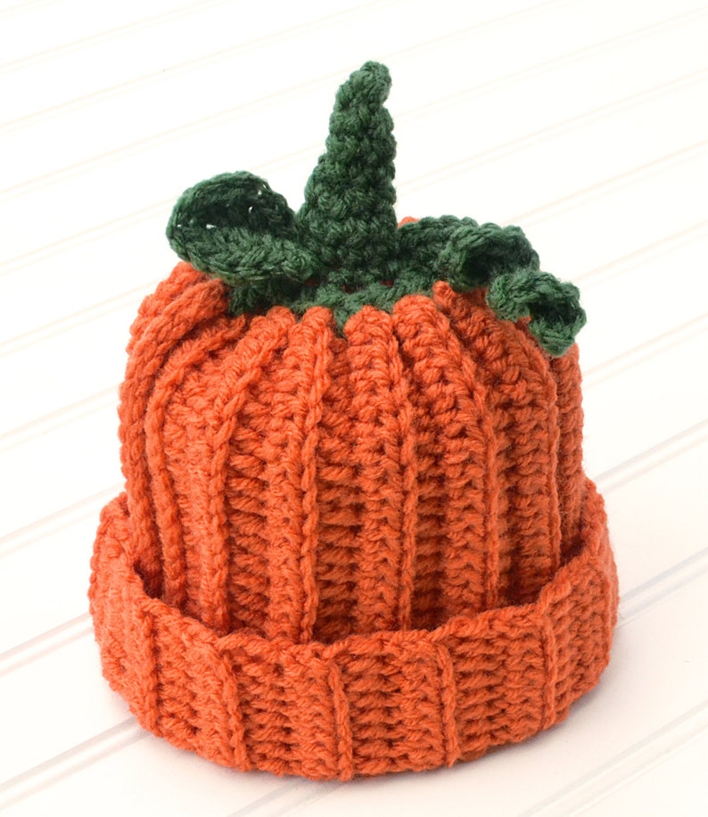 Op de afbeelding: Een gebreide pompoenmuts met een groene steel en bladeren. De muts is oranje en heeft een geribbelde textuur.