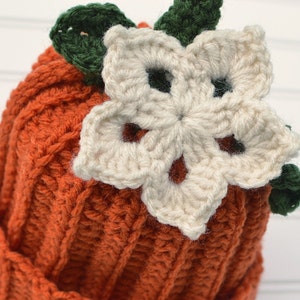 Op de afbeelding: Een gebreide pompoenmuts met een witte bloem en groene bladeren erop. De muts is oranje en de bloem is cr&egrave;mekleurig.