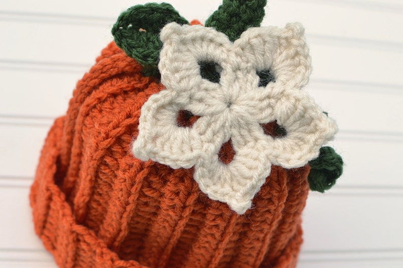 Op de afbeelding: Een gebreide pompoenmuts met een witte bloem en groene bladeren erop. De muts is oranje en de bloem is cr&egrave;mekleurig.