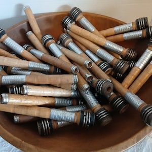 May include: A wooden bowl filled with a large quantity of wooden spools with silver and black metal accents. The spools are used for winding yarn.