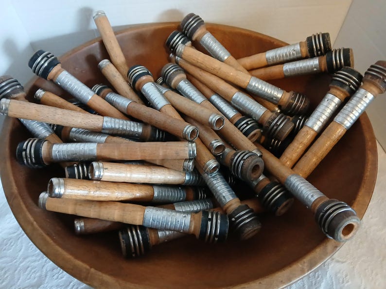 May include: A wooden bowl filled with a large quantity of wooden spools with silver and black metal accents. The spools are used for winding yarn.