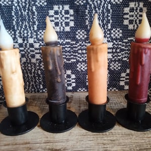May include: Four taper candles in different colors, including beige, brown, orange, and red, are displayed on black candle holders. The candles are dripped and have a realistic flame effect.