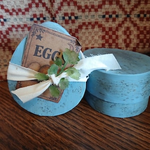 May include: A light blue, oval-shaped wooden box with a matching lid. The box has a tag that reads "EGGS" and is decorated with a white ribbon and faux greenery. The box is sitting on a wooden surface.