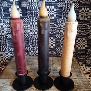 May include: Three pillar candles with black metal candle holders. The candles are red, black, and cream colored.