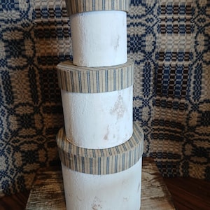 Puede incluir: Tres cajas cilíndricas blancas apiladas. Cada caja tiene una tapa con una banda de tela a rayas azules y beige. Las cajas están sobre una superficie de madera, con un fondo de tela estampada.