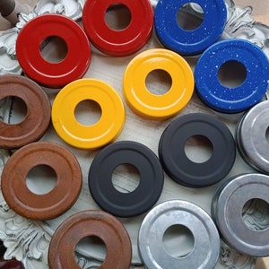 May include: A collection of colorful jar lids in various shades, including red, yellow, blue with white speckles, brown, black, and silver. The lids are arranged on a decorative, white-framed tray, showcasing their circular design and central hole.