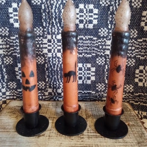 May include: Three Halloween themed candles with black metal candle holders. The candles are orange with black designs. One candle has a jack-o-lantern face, one has a black cat, and one has spiders.
