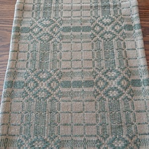 May include: A rectangular, woven table runner with a repeating geometric pattern in shades of green and tan. The design features squares, diamonds, and other shapes. The runner is made of a textured fabric and is suitable for a dining table or other surface.