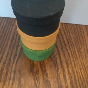May include: A stack of three cylindrical containers with lids. The top container is black, the middle container is orange, and the bottom container is green. Each container features a smooth, matte finish and a simple, uniform design with a circular lid.