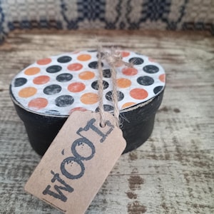May include: A small, black, oval-shaped box with a lid decorated with orange, black, and white polka dots. A brown tag with the word "WOOI" is attached with twine. The box is on a wooden surface.