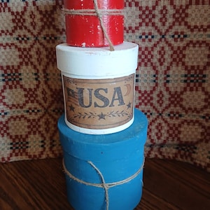 May include: Three stacked, round, painted boxes in red, white, and blue. The middle box has a label with "USA" and stars. The boxes are tied with twine. The theme is patriotic and decorative.