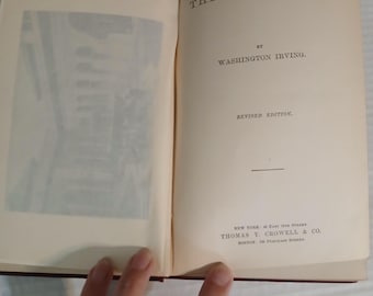 May include: Open book titled "THE ALHAMBRA" by Washington Irving. The book is a revised edition, with text printed in black on aged, cream-colored pages. The cover is a dark burgundy color. The book is open, revealing an illustration on the left page.