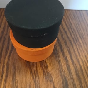 May include: A cylindrical container with a matte black lid stacked on top of a bright orange base. The object features a simple, two-tone design with a flat circular top and smooth, solid-colored sides, creating a distinct contrast between the black and orange sections.