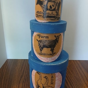 May include: A stack of three cylindrical blue boxes with distressed edges. Each box features a tan oval label with a black illustration: a cow with the text Fresh Milk, a goat with the word Farm, and two geese with the vertical text FARM.