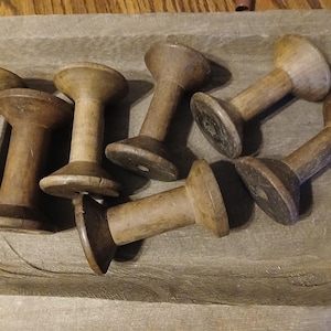 May include: A collection of seven antique wooden spools in varying shades of brown. The spools are cylindrical with a flared top and bottom, resting on a weathered, gray wooden surface. The spools are arranged in a slightly haphazard manner.