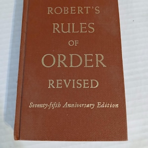 May include: A brown book titled "ROBERT'S RULES OF ORDER REVISED" with gold lettering. The cover also includes "Seventy-fifth Anniversary Edition". The book is closed and slightly worn.