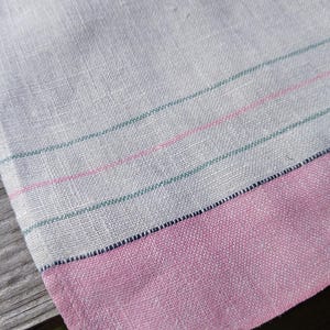 May include: A light gray linen towel with a pink border. The towel has three horizontal stripes in pink and green. The fabric has a woven texture, and the border is a solid pink color.